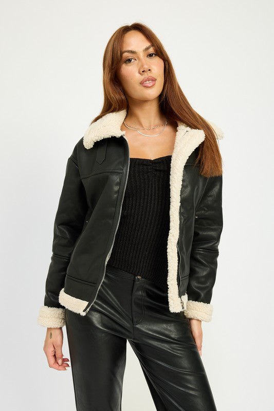 A person with long hair wearing the SHEARLING MOTO JACKET, featuring a black leather exterior, shearling collar, and polyester lining, along with black pants, poses against a plain background.