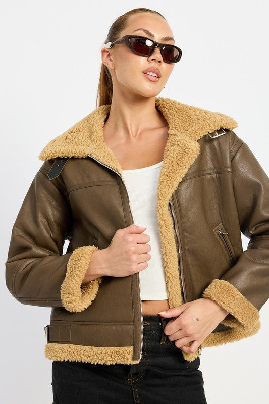 A person with long hair wearing the SHEARLING MOTO JACKET, featuring a black leather exterior, shearling collar, and polyester lining, along with black pants, poses against a plain background.