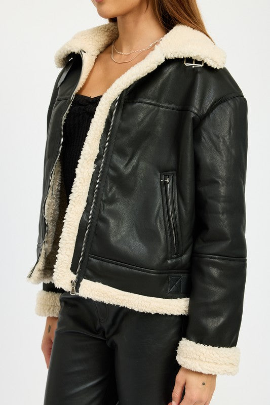 A person with long hair wearing the SHEARLING MOTO JACKET, featuring a black leather exterior, shearling collar, and polyester lining, along with black pants, poses against a plain background.