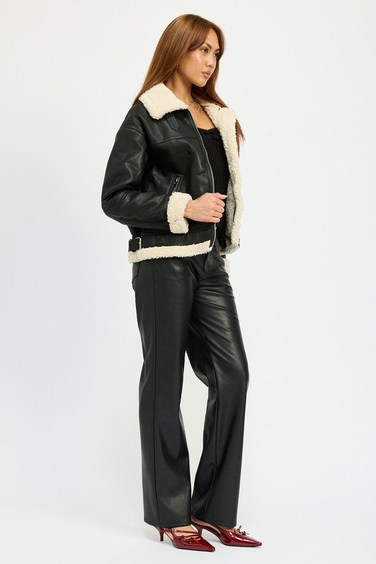 A person with long hair wearing the SHEARLING MOTO JACKET, featuring a black leather exterior, shearling collar, and polyester lining, along with black pants, poses against a plain background.