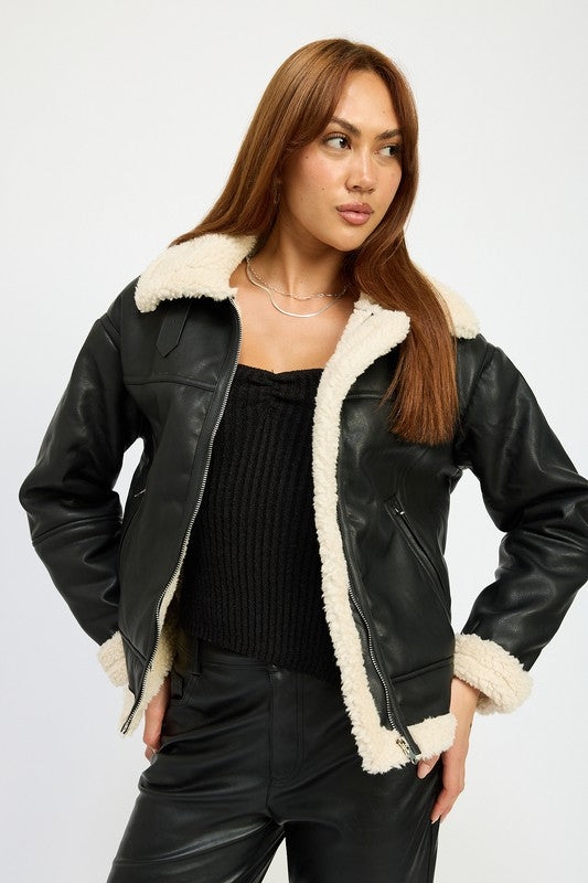 A person with long hair wearing the SHEARLING MOTO JACKET, featuring a black leather exterior, shearling collar, and polyester lining, along with black pants, poses against a plain background.