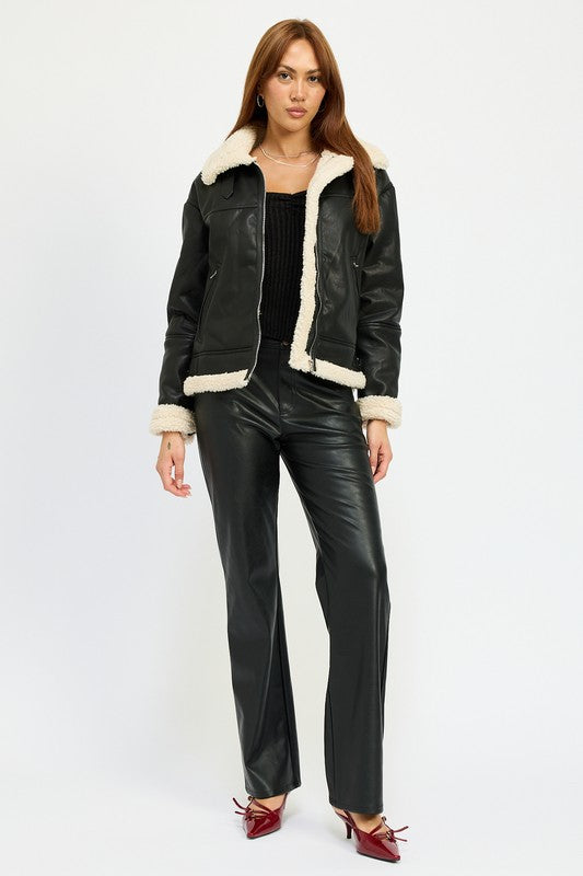 A person with long hair wearing the SHEARLING MOTO JACKET, featuring a black leather exterior, shearling collar, and polyester lining, along with black pants, poses against a plain background.