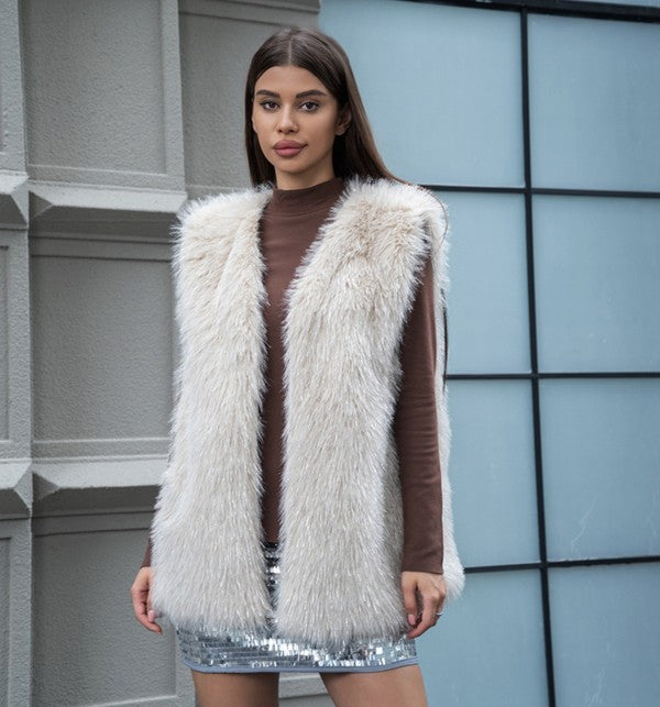 A woman stands in front of a building with a geometric wall, wearing a Long Fax Fur Cardigan in light blue over a long-sleeved white top and a black sequin skirt.