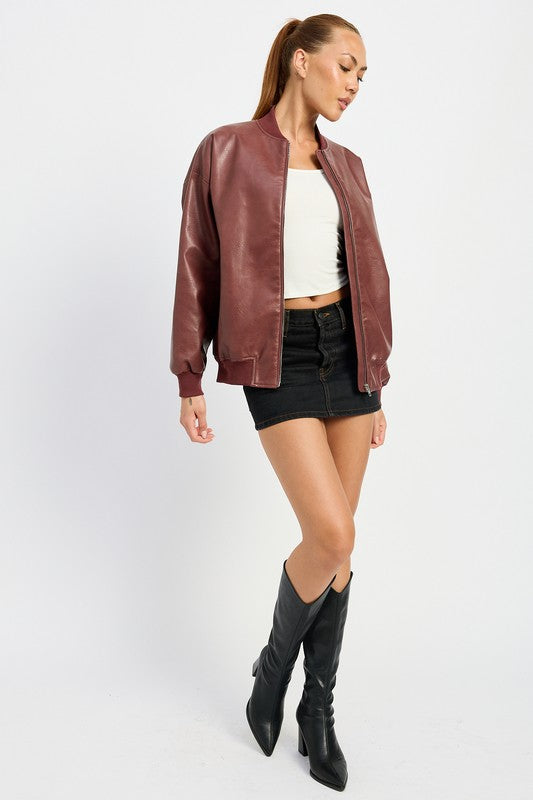 A person poses wearing a VINTAGE LEATHER BOMBER JACKET in maroon, made of polyurethane, over a white crop top and a black mini skirt, standing with hands in jacket pockets against a plain white background.