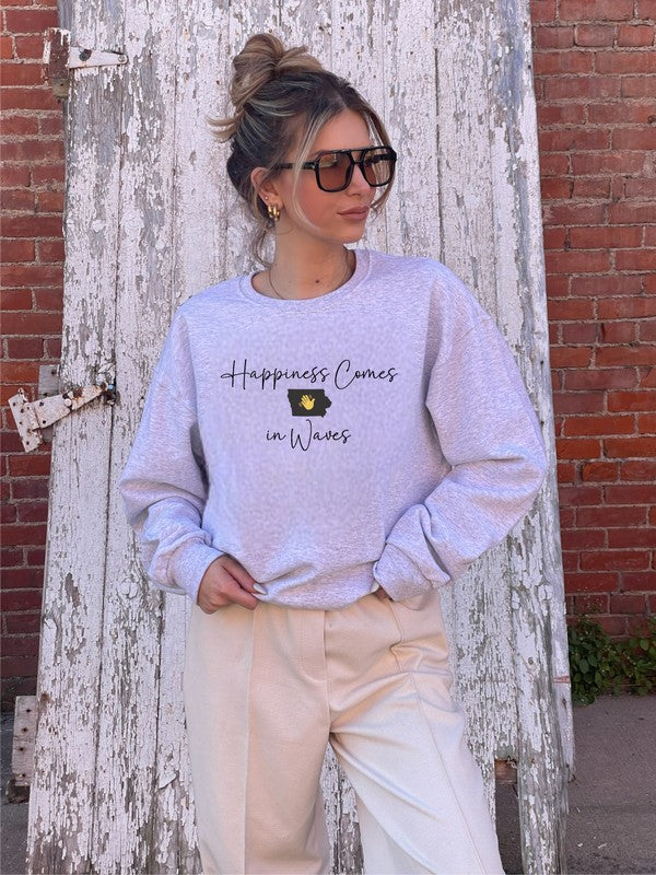 A person wearing the "Happiness Comes in Waves Cozy Sweatshirt," which features an illustration of a wave and a sun along with the text, effortlessly combines comfort and style. The unisex garment pairs perfectly with the person's long hair and orange-painted nails.