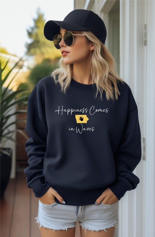 A person wearing the "Happiness Comes in Waves Cozy Sweatshirt," which features an illustration of a wave and a sun along with the text, effortlessly combines comfort and style. The unisex garment pairs perfectly with the person's long hair and orange-painted nails.