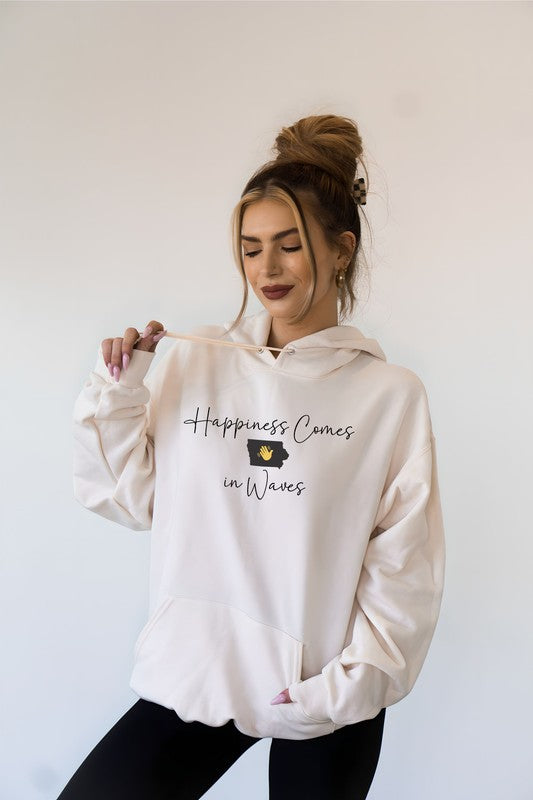 A person in a black Happiness Comes in Waves Hoodie Sweatshirt holds a coffee cup while smiling outdoors.