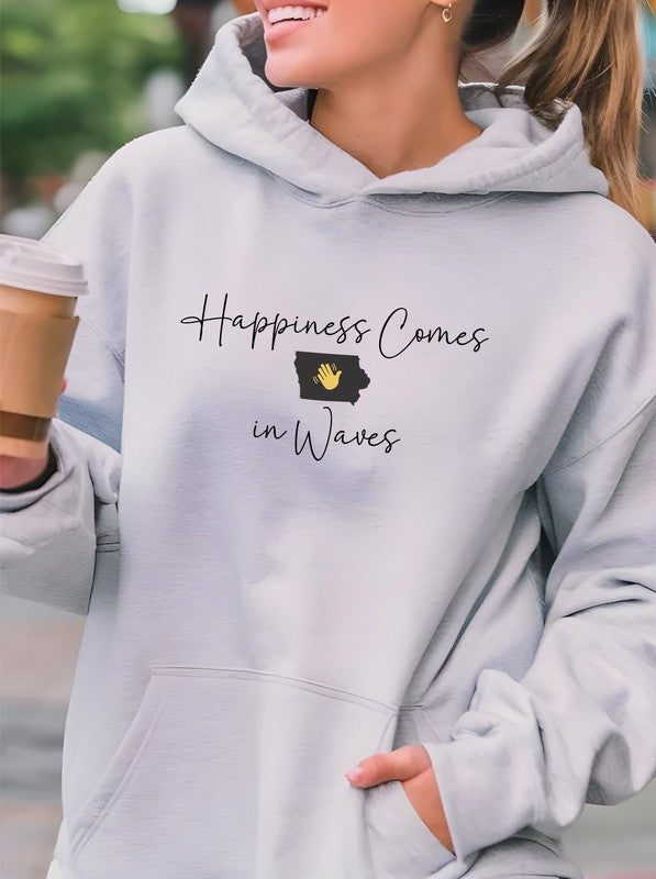 A person in a black Happiness Comes in Waves Hoodie Sweatshirt holds a coffee cup while smiling outdoors.