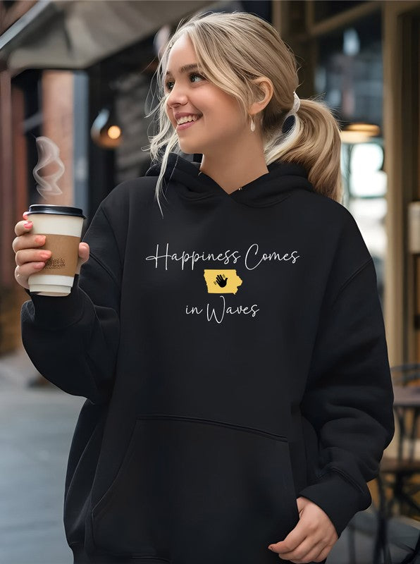 A person in a black Happiness Comes in Waves Hoodie Sweatshirt holds a coffee cup while smiling outdoors.