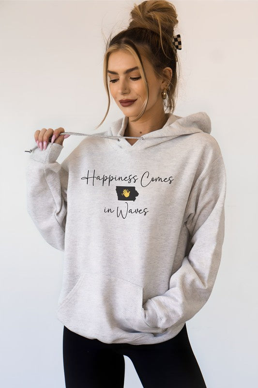 A person in a black Happiness Comes in Waves Hoodie Sweatshirt holds a coffee cup while smiling outdoors.