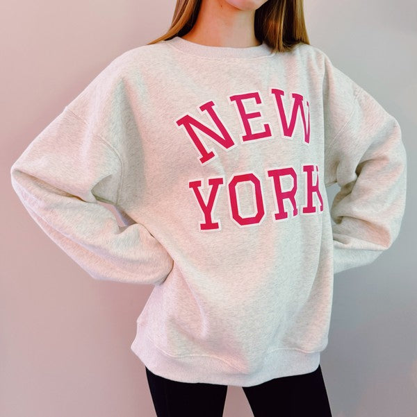 A person stands outside on grass, wearing an oversized New York Sweatshirt in quality materials with "New York" written in red letters, and black pants. Both arms are raised and outstretched. Trees are visible in the background.