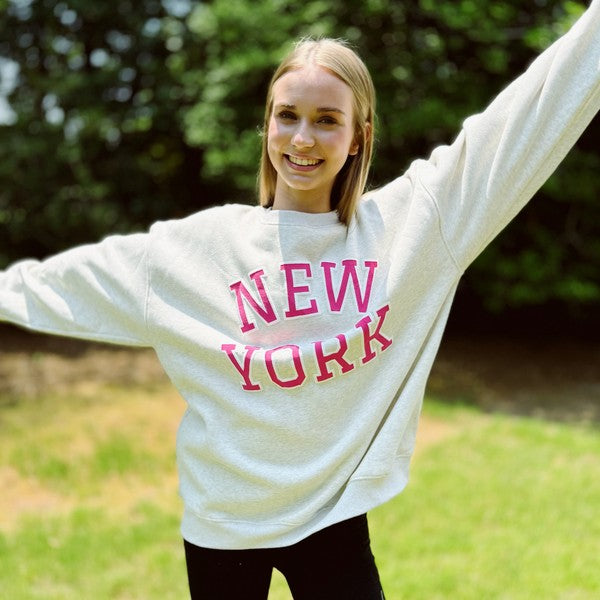 A person stands outside on grass, wearing an oversized New York Sweatshirt in quality materials with "New York" written in red letters, and black pants. Both arms are raised and outstretched. Trees are visible in the background.