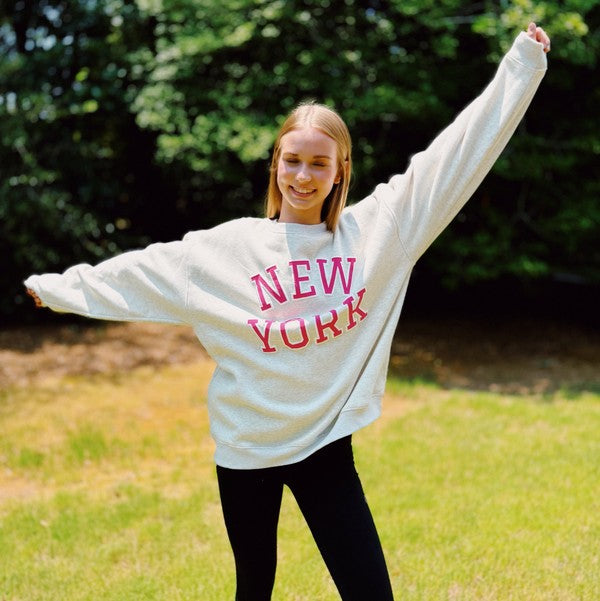 A person stands outside on grass, wearing an oversized New York Sweatshirt in quality materials with "New York" written in red letters, and black pants. Both arms are raised and outstretched. Trees are visible in the background.