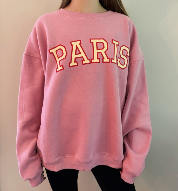 A person wearing a comfortable and stylish pink Paris Sweatshirt, a true wardrobe essential, sits on a white couch with arms resting on the backrest.