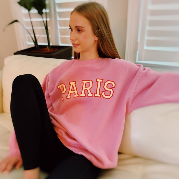 A person wearing a comfortable and stylish pink Paris Sweatshirt, a true wardrobe essential, sits on a white couch with arms resting on the backrest.