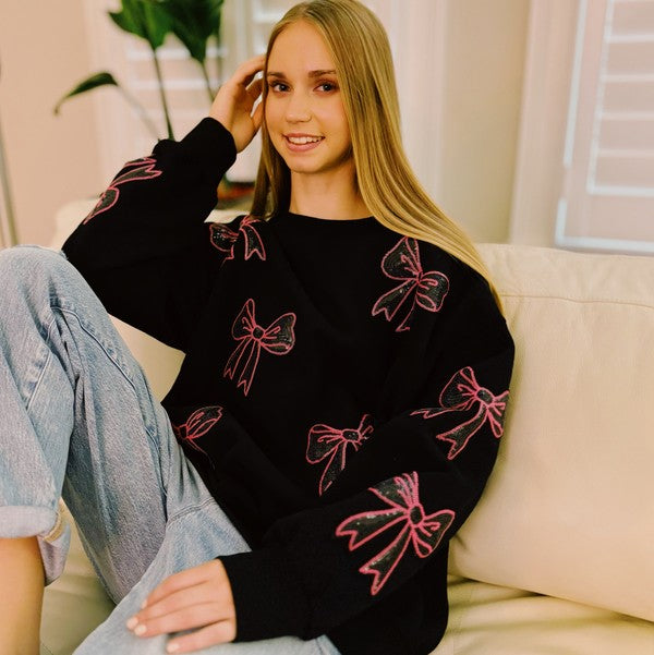 A young woman with long blonde hair sits on a white couch, wearing a cozy and chic Sparkly Bow Patch Sweatshirt paired with light blue jeans. She smiles at the camera.