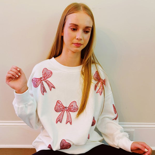 A young woman with long blonde hair sits on a white couch, wearing a cozy and chic Sparkly Bow Patch Sweatshirt paired with light blue jeans. She smiles at the camera.