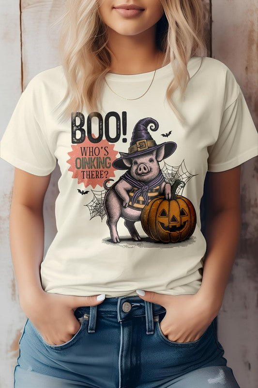 A woman wears the Boo Who's Oinking There Halloween Graphic Tee, featuring a cartoon pig in a witch hat alongside a pumpkin and the playful phrase "Boo! Who's Oinking There?" This unisex top perfectly blends style and comfort.