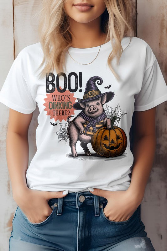 A woman wears the Boo Who's Oinking There Halloween Graphic Tee, featuring a cartoon pig in a witch hat alongside a pumpkin and the playful phrase "Boo! Who's Oinking There?" This unisex top perfectly blends style and comfort.