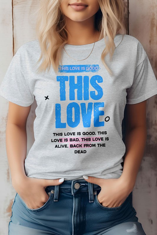 A person wearing the "This Love, Music Graphic Tee" by Bella Canvas, which features blue and black text inspired by Taylor Swift lyrics that reads "This Love is Good. This Love is Bad. This Love is Alive. Back from the Dead," stands with hands in their jeans pockets.