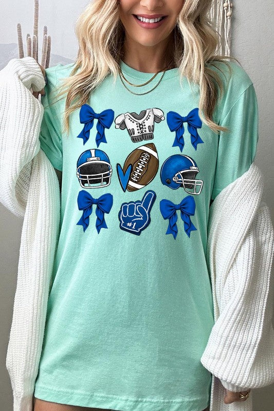 A woman wearing a Blue Coquette Football Graphic Tee adorned with blue bows, helmets, a football, a foam finger, and a jersey. She has yellow painted nails and is holding her shirt. This high-quality printed design guarantees long-lasting wear and vibrant colors.