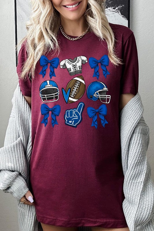 A woman wearing a Blue Coquette Football Graphic Tee adorned with blue bows, helmets, a football, a foam finger, and a jersey. She has yellow painted nails and is holding her shirt. This high-quality printed design guarantees long-lasting wear and vibrant colors.