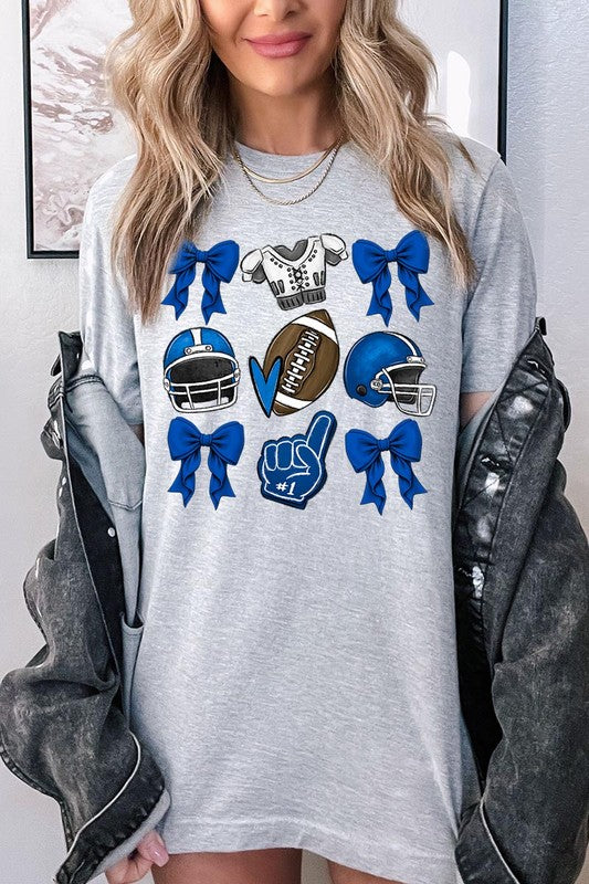 A woman wearing a Blue Coquette Football Graphic Tee adorned with blue bows, helmets, a football, a foam finger, and a jersey. She has yellow painted nails and is holding her shirt. This high-quality printed design guarantees long-lasting wear and vibrant colors.