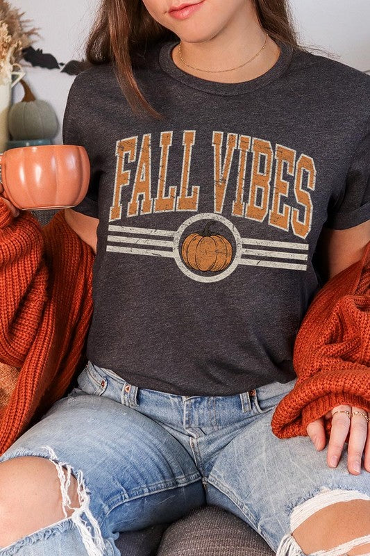 A woman wearing a wide-brimmed hat and a yellow Fall Vibes Graphic Tee featuring a pumpkin graphic stands smiling while holding a light-colored jacket.