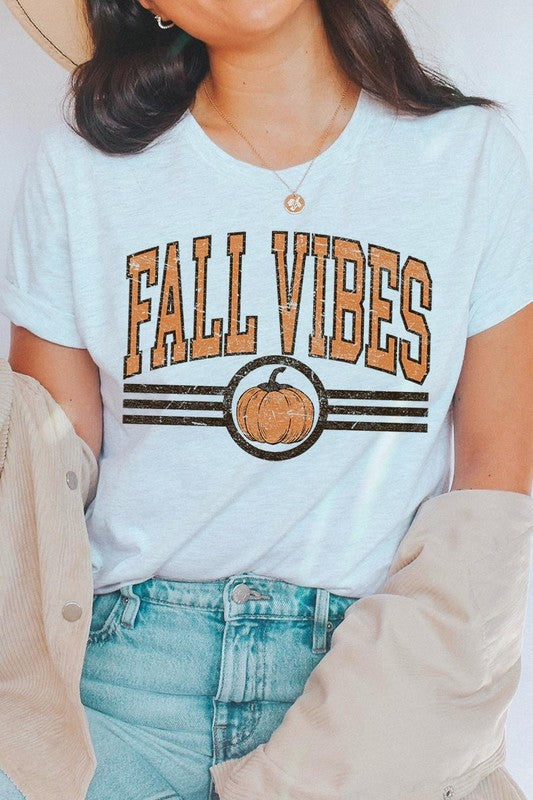 A woman wearing a wide-brimmed hat and a yellow Fall Vibes Graphic Tee featuring a pumpkin graphic stands smiling while holding a light-colored jacket.