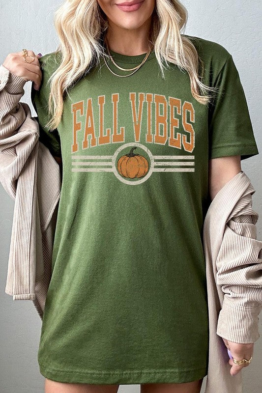 A woman wearing a wide-brimmed hat and a yellow Fall Vibes Graphic Tee featuring a pumpkin graphic stands smiling while holding a light-colored jacket.