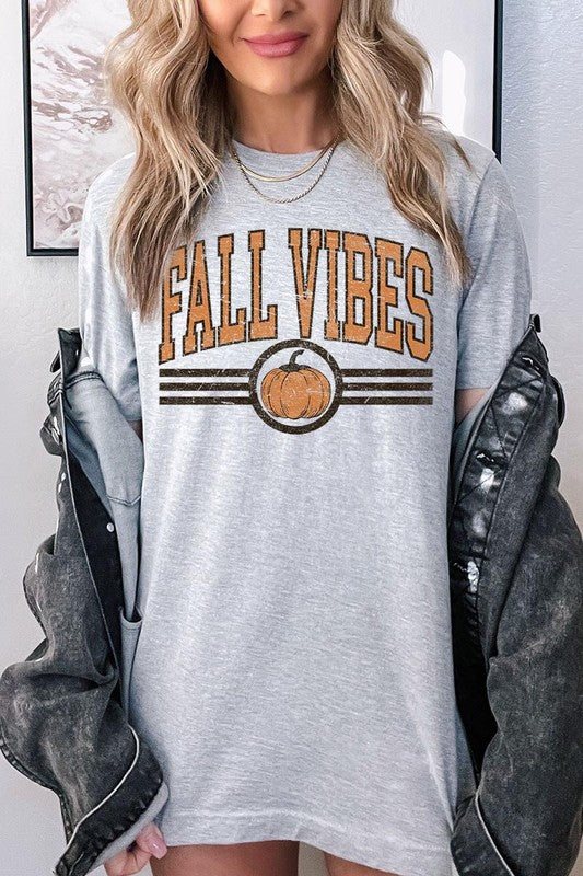 A woman wearing a wide-brimmed hat and a yellow Fall Vibes Graphic Tee featuring a pumpkin graphic stands smiling while holding a light-colored jacket.