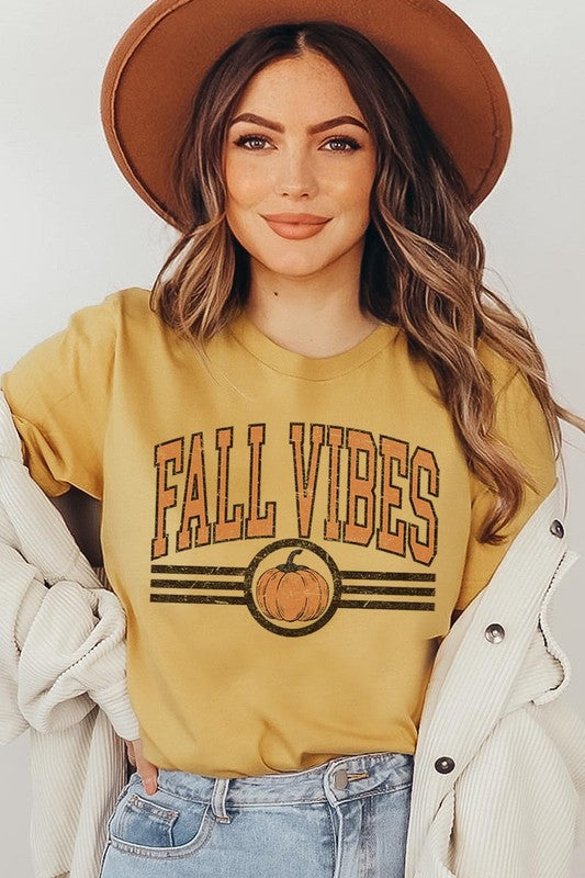 A woman wearing a wide-brimmed hat and a yellow Fall Vibes Graphic Tee featuring a pumpkin graphic stands smiling while holding a light-colored jacket.