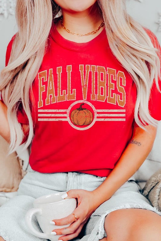 A woman wearing a wide-brimmed hat and a yellow Fall Vibes Graphic Tee featuring a pumpkin graphic stands smiling while holding a light-colored jacket.