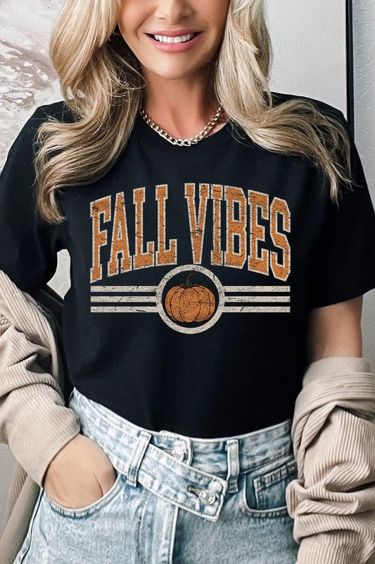 A woman wearing a wide-brimmed hat and a yellow Fall Vibes Graphic Tee featuring a pumpkin graphic stands smiling while holding a light-colored jacket.