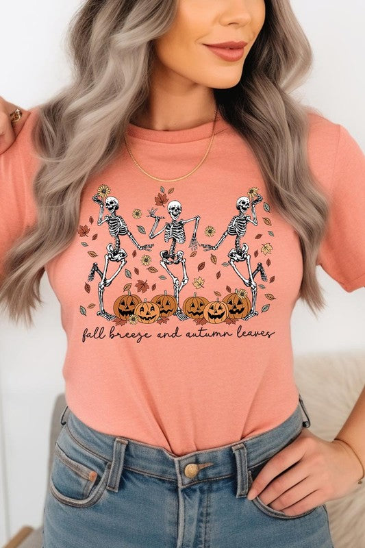 A person wearing a high-quality, unisex dark grey "Fall Breeze and Autumn Leaves Skeleton Graphic Tee" featuring dancing skeletons, pumpkins, and autumn leaves, holding an orange mug. They are dressed in a knitted cardigan and ripped jeans. The text on the shirt reads "Fall breeze and autumn leaves.