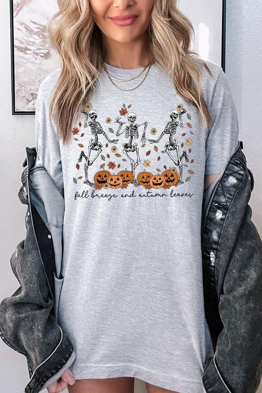 A person wearing a high-quality, unisex dark grey "Fall Breeze and Autumn Leaves Skeleton Graphic Tee" featuring dancing skeletons, pumpkins, and autumn leaves, holding an orange mug. They are dressed in a knitted cardigan and ripped jeans. The text on the shirt reads "Fall breeze and autumn leaves.