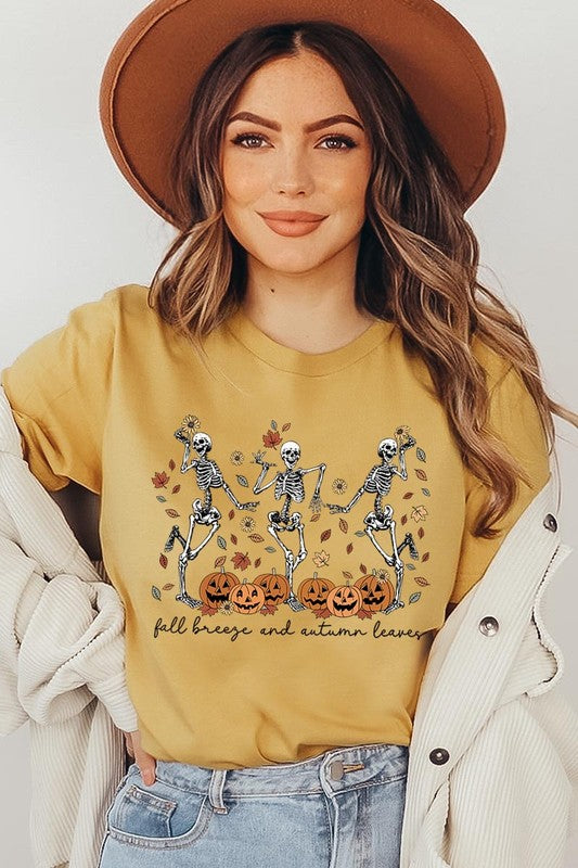 A person wearing a high-quality, unisex dark grey "Fall Breeze and Autumn Leaves Skeleton Graphic Tee" featuring dancing skeletons, pumpkins, and autumn leaves, holding an orange mug. They are dressed in a knitted cardigan and ripped jeans. The text on the shirt reads "Fall breeze and autumn leaves.