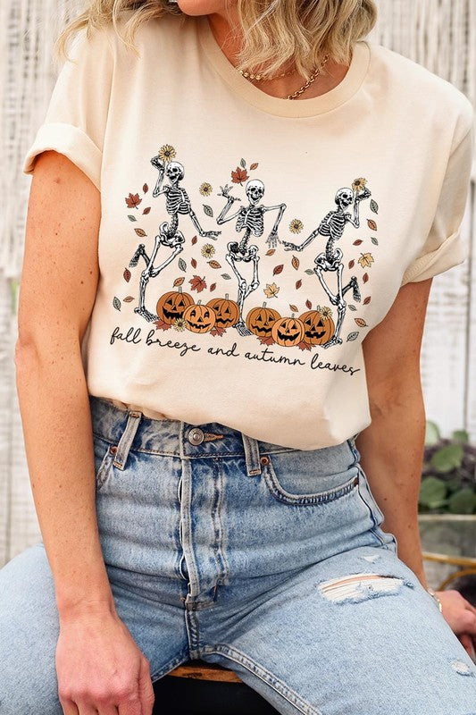 A person wearing a high-quality, unisex dark grey "Fall Breeze and Autumn Leaves Skeleton Graphic Tee" featuring dancing skeletons, pumpkins, and autumn leaves, holding an orange mug. They are dressed in a knitted cardigan and ripped jeans. The text on the shirt reads "Fall breeze and autumn leaves.