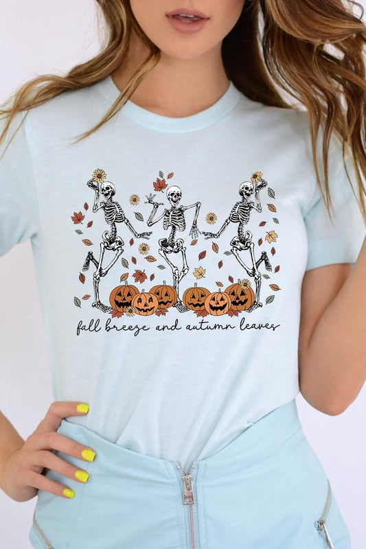 A person wearing a high-quality, unisex dark grey "Fall Breeze and Autumn Leaves Skeleton Graphic Tee" featuring dancing skeletons, pumpkins, and autumn leaves, holding an orange mug. They are dressed in a knitted cardigan and ripped jeans. The text on the shirt reads "Fall breeze and autumn leaves.