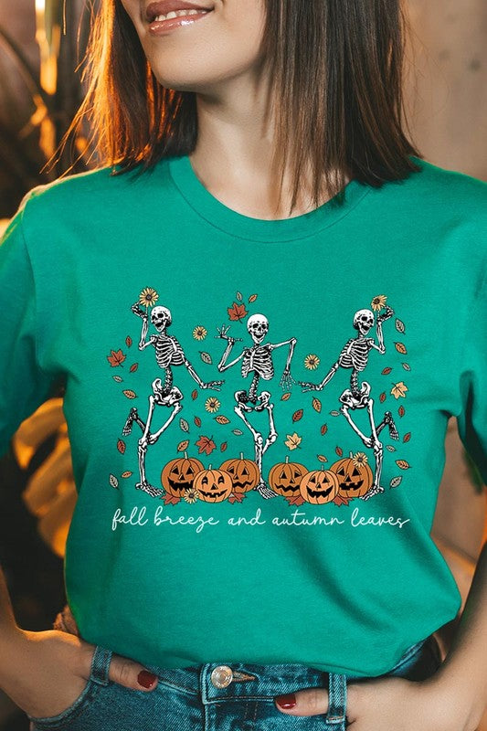 A person wearing a high-quality, unisex dark grey "Fall Breeze and Autumn Leaves Skeleton Graphic Tee" featuring dancing skeletons, pumpkins, and autumn leaves, holding an orange mug. They are dressed in a knitted cardigan and ripped jeans. The text on the shirt reads "Fall breeze and autumn leaves.