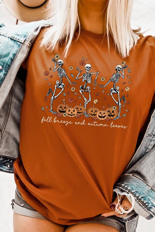 A person wearing a high-quality, unisex dark grey "Fall Breeze and Autumn Leaves Skeleton Graphic Tee" featuring dancing skeletons, pumpkins, and autumn leaves, holding an orange mug. They are dressed in a knitted cardigan and ripped jeans. The text on the shirt reads "Fall breeze and autumn leaves.