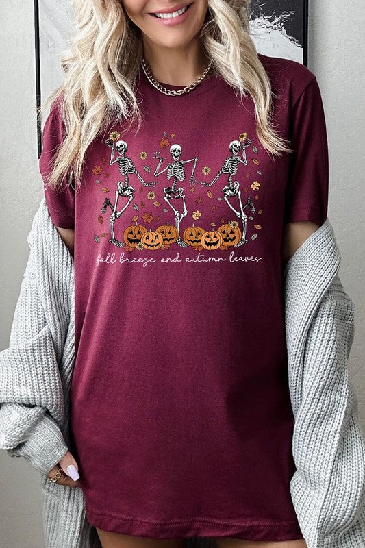 A person wearing a high-quality, unisex dark grey "Fall Breeze and Autumn Leaves Skeleton Graphic Tee" featuring dancing skeletons, pumpkins, and autumn leaves, holding an orange mug. They are dressed in a knitted cardigan and ripped jeans. The text on the shirt reads "Fall breeze and autumn leaves.