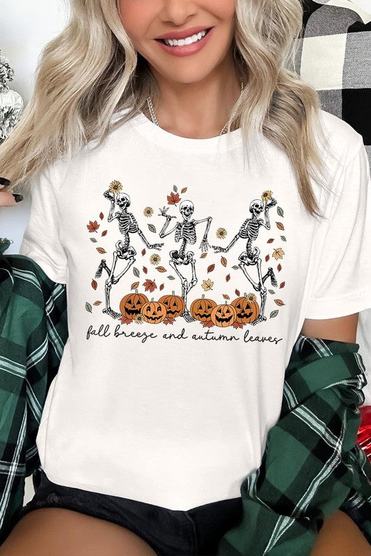 A person wearing a high-quality, unisex dark grey "Fall Breeze and Autumn Leaves Skeleton Graphic Tee" featuring dancing skeletons, pumpkins, and autumn leaves, holding an orange mug. They are dressed in a knitted cardigan and ripped jeans. The text on the shirt reads "Fall breeze and autumn leaves.