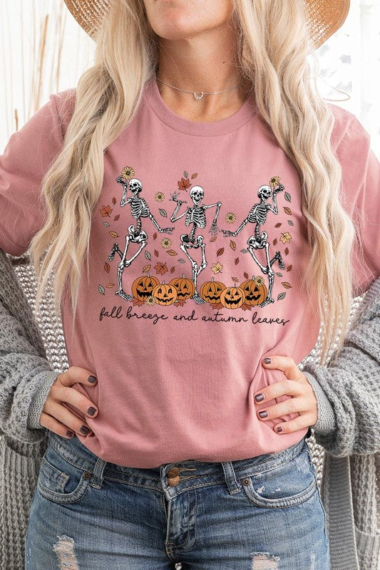 A person wearing a high-quality, unisex dark grey "Fall Breeze and Autumn Leaves Skeleton Graphic Tee" featuring dancing skeletons, pumpkins, and autumn leaves, holding an orange mug. They are dressed in a knitted cardigan and ripped jeans. The text on the shirt reads "Fall breeze and autumn leaves.