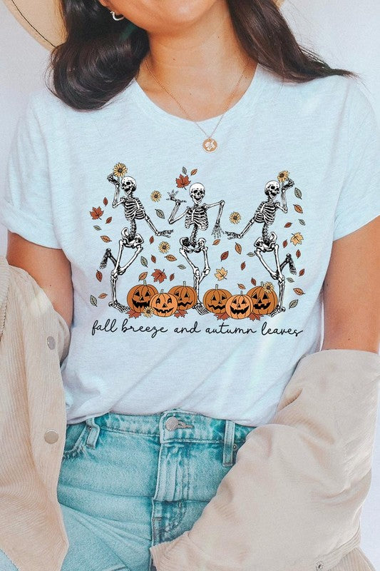 A person wearing a high-quality, unisex dark grey "Fall Breeze and Autumn Leaves Skeleton Graphic Tee" featuring dancing skeletons, pumpkins, and autumn leaves, holding an orange mug. They are dressed in a knitted cardigan and ripped jeans. The text on the shirt reads "Fall breeze and autumn leaves.