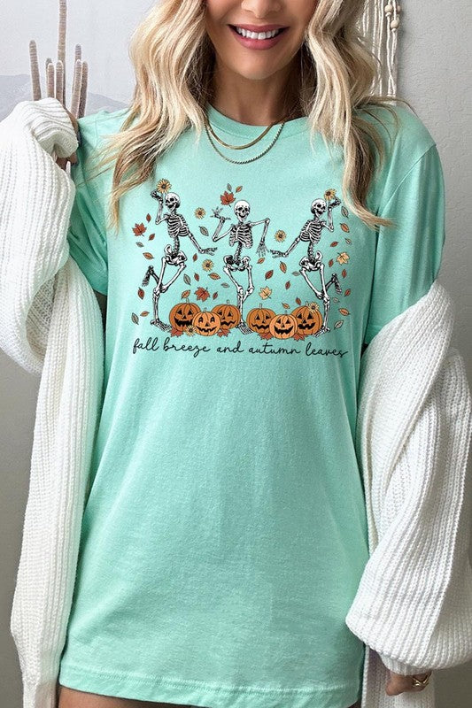 A person wearing a high-quality, unisex dark grey "Fall Breeze and Autumn Leaves Skeleton Graphic Tee" featuring dancing skeletons, pumpkins, and autumn leaves, holding an orange mug. They are dressed in a knitted cardigan and ripped jeans. The text on the shirt reads "Fall breeze and autumn leaves.