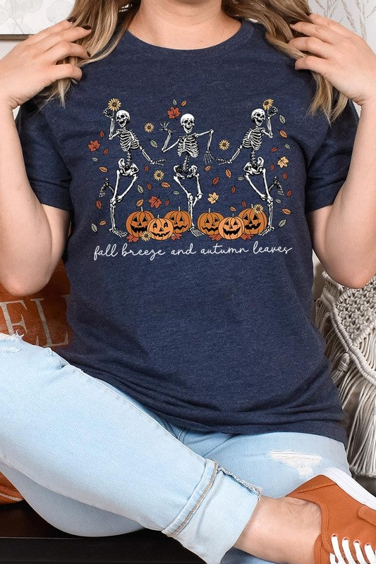 A person wearing a high-quality, unisex dark grey "Fall Breeze and Autumn Leaves Skeleton Graphic Tee" featuring dancing skeletons, pumpkins, and autumn leaves, holding an orange mug. They are dressed in a knitted cardigan and ripped jeans. The text on the shirt reads "Fall breeze and autumn leaves.