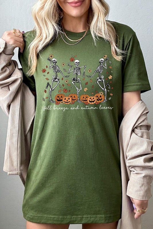 A person wearing a high-quality, unisex dark grey "Fall Breeze and Autumn Leaves Skeleton Graphic Tee" featuring dancing skeletons, pumpkins, and autumn leaves, holding an orange mug. They are dressed in a knitted cardigan and ripped jeans. The text on the shirt reads "Fall breeze and autumn leaves.