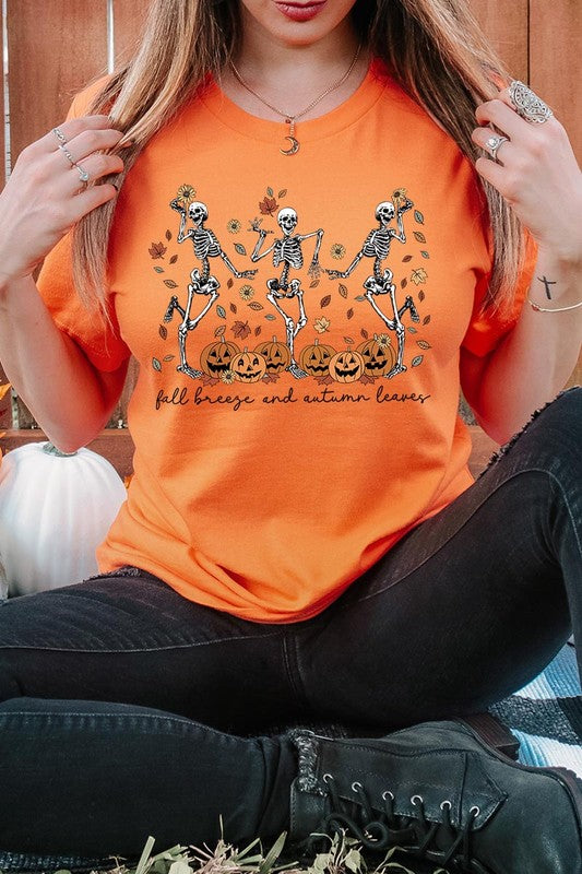 A person wearing a high-quality, unisex dark grey "Fall Breeze and Autumn Leaves Skeleton Graphic Tee" featuring dancing skeletons, pumpkins, and autumn leaves, holding an orange mug. They are dressed in a knitted cardigan and ripped jeans. The text on the shirt reads "Fall breeze and autumn leaves.