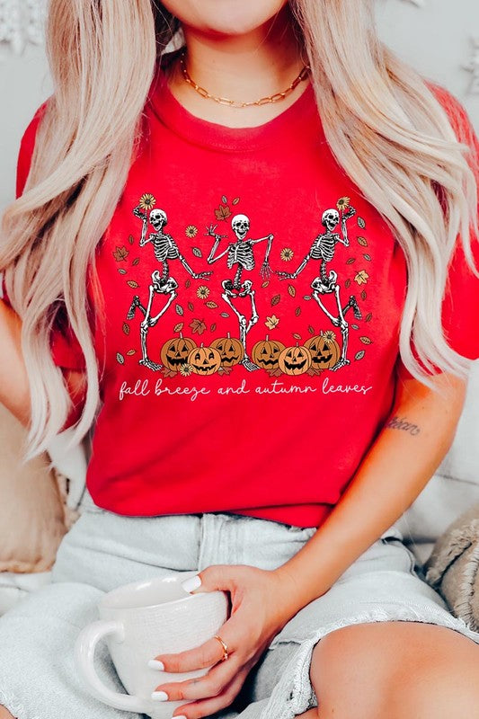 A person wearing a high-quality, unisex dark grey "Fall Breeze and Autumn Leaves Skeleton Graphic Tee" featuring dancing skeletons, pumpkins, and autumn leaves, holding an orange mug. They are dressed in a knitted cardigan and ripped jeans. The text on the shirt reads "Fall breeze and autumn leaves.